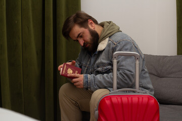 Travel photo. Sad mature man with beard, blue denim jeans jacket sits with red suitcase on sofa in...
