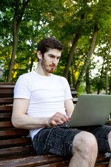 Young man in white t-shirt working on laptop in park. Side view of guy typing on bench outdoors.