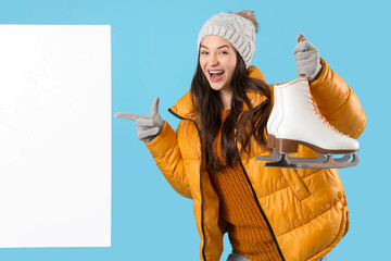 Young woman in winter clothes with ice skates pointing at blank poster on blue background