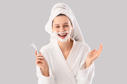 Beautiful young happy woman in bathrobe with razor and shaving foam on grey background - Powered by Adobe