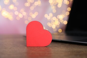 Online dating site. Decorative heart and laptop on wooden table against blurred lights, closeup....