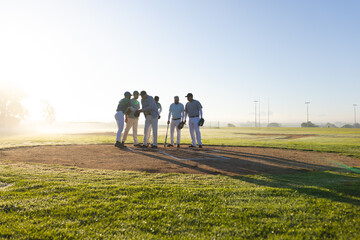 Diverse male teammates huddling on baseball mound at sunrise, holding bat and reviewing tablet