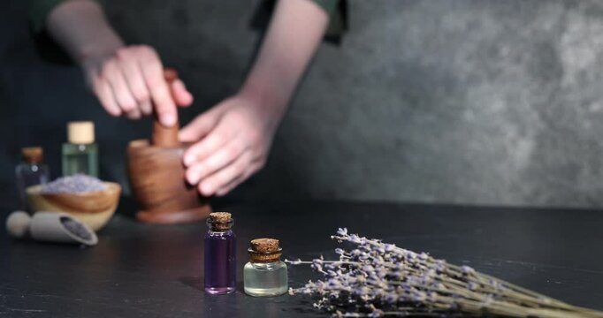 Woman using mortar and pestle at black table, focus on lavender flowers and bottles of essential oil