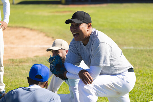 Diverse male teammates kneeling on baseball field near infield, wearing caps and holding gloves