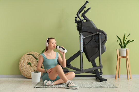 Sporty young woman with headphones drinking water near modern exercising bike at home