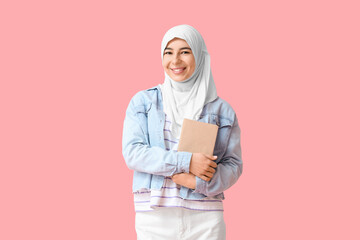 Beautiful young Muslim woman in hijab with book on pink background