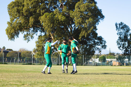 Three male baseball teammates celebrating, tossing glove, wearing caps, cleats on field near fence