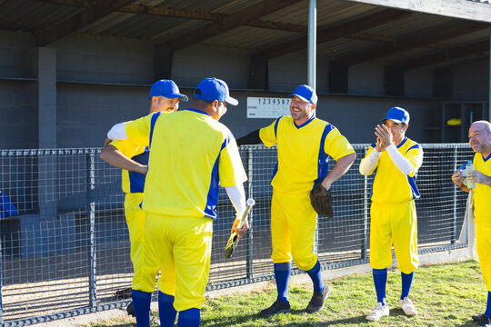 Male baseball teammates huddling beside dugout fence at baseball field holding bat and glove