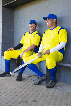 Two male baseball teammates sitting on bench in dugout leaning against wall holding glove and bat