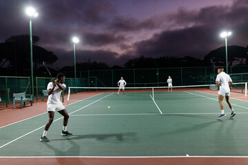 Diverse male tennis players playing doubles on court at dusk under lights with rackets, ball, net
