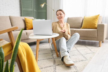 Smiling young woman using laptop on table near grey sofa in living room