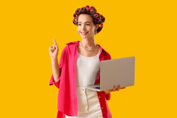 Young woman with hair curlers and laptop pointing at something on yellow background