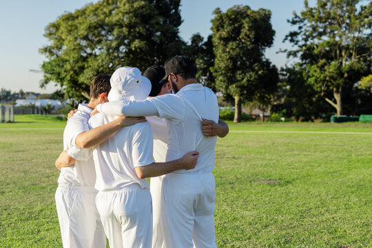 Diverse male teammates huddling on grassy cricket field discussing strategy with white uniforms