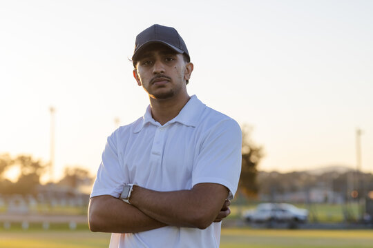 Indian man standing with arms crossed on sports field at sunset wearing smartwatch and baseball cap
