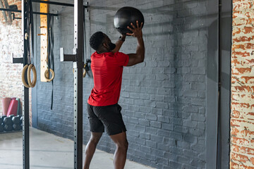 African American man in red t-shirt, black shorts tossing medicine ball against gym wall with rings