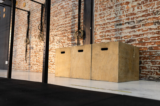 Wooden plyometric boxes resting on white gym floor against brick wall beside rig with hanging rings