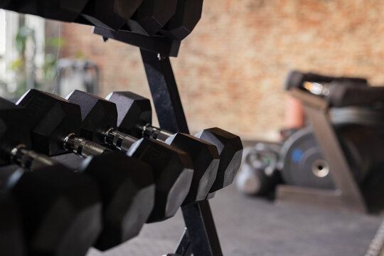 Black hexagonal dumbbells are resting on metal rack in gym near treadmill with natural light