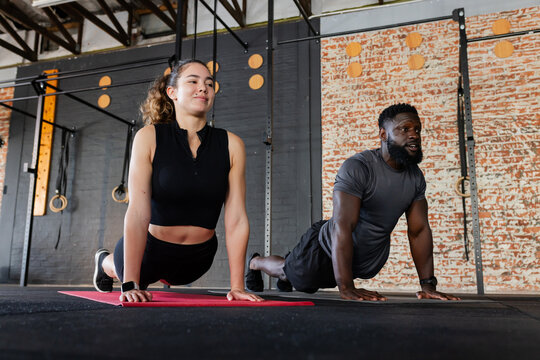 Diverse workout partners performing cobra stretches on yoga mats in fitness studio with pull-up rig