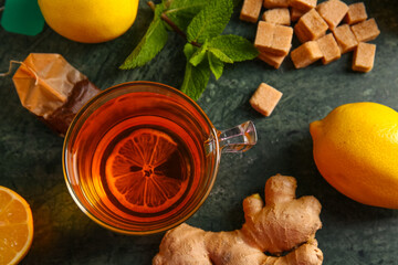 Cup of hot beverage with tea bag, lemons, ginger, mint leaves and sugar cubes on green grunge...