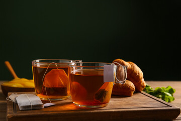 Cups of hot beverage with tea bags, croissants and mint leaves on wooden table against dark wall