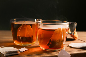 Cups of hot beverage with tea bags on wooden table against dark wall