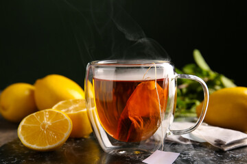 Cup of hot beverage with tea bags, mint leaves and lemons on board against dark wall