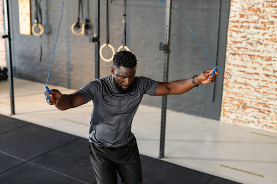 African American man skipping with blue jump rope in gym with pull-up rig and rings