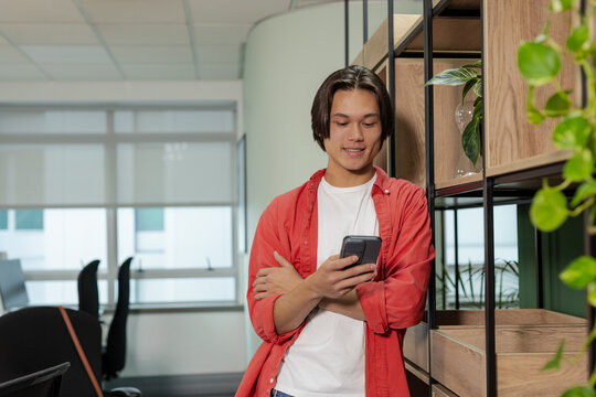Asian man leaning on shelving with plants, checking smartphone in office with chairs, roller blinds