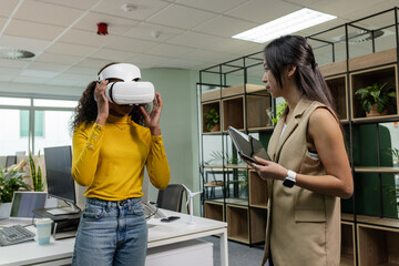 Diverse coworkers adjusting VR headset and observing tablet at open-plan office desk with monitors