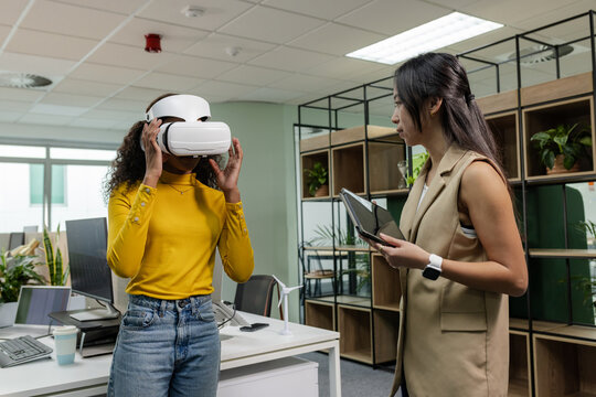 Diverse coworkers adjusting VR headset and observing tablet at open-plan office desk with monitors