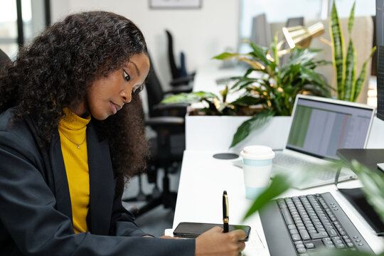 African American woman taking notes in notebook at desk with laptop in open-plan office, copy space