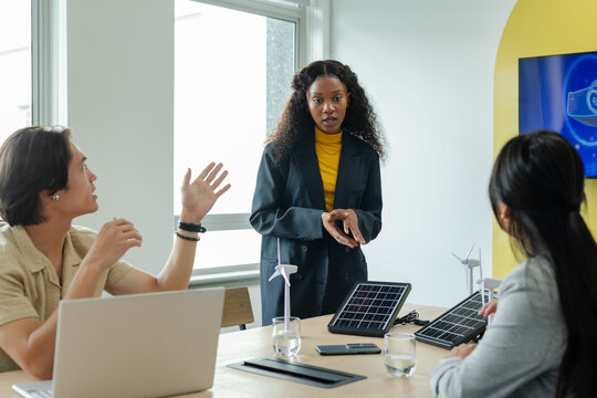 Diverse coworkers in business attire examining solar panels, wind turbines, laptop at meeting table