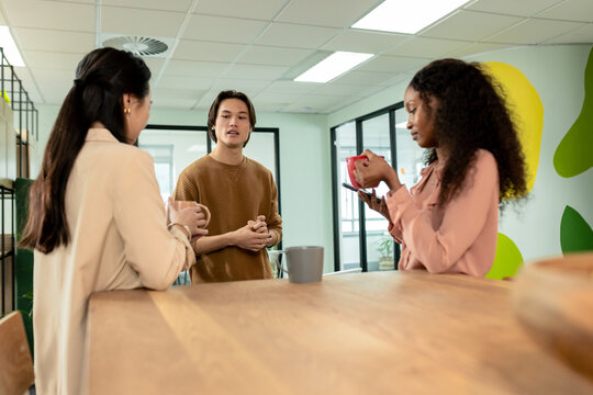 Diverse coworkers standing around wooden table at office break area holding coffee mugs and phone