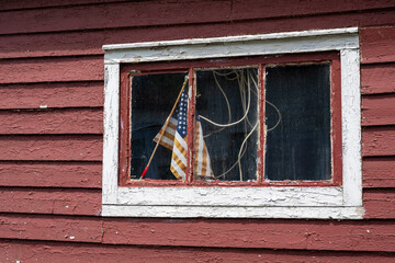 An old weathered and worn American flag hangs on a small wooden pole in the window of a vintage red wood shed with white trim. The cloth flag is tattered and the sun has faded and weakened the fabric 