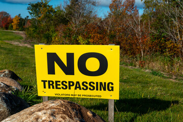 A yellow warning sign with black lettering. The text spells out no trespassing violators will be prosecuted. The private property signpost is on a sandy beach with grass, large rocks, and a blue ocean