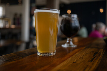 A tall, cold glass of hazy IPA beer with froth on top. The clear pint glass of ale is on a brown table with a metal fence in the background. The liquid is a bright yellow color with the sun is shining
