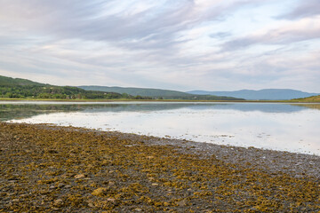 A seascape with wispy blue, pink, and pale yellow clouds. The calm, freshwater reflects the...