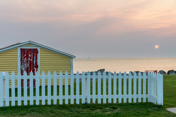 A bright yellow shed with a wooden red door on the edge of a cliff overlooking the ocean. The sky is pale pink with a setting sun. There's an iceberg in the distance. The ground is covered with grass.