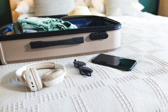 Open suitcase lying on hotel bed with folded clothes, straw hat, headphones, sunglasses, smartphone