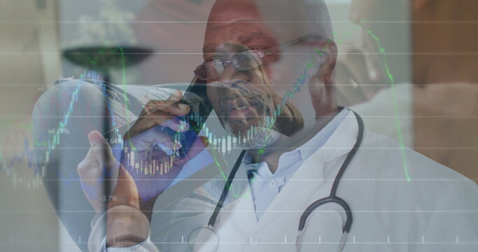 Speaking senior physician in white coat holding smartphone at clinic, stethoscope and market charts