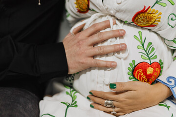 Close-up of couple’s hands on pregnant belly with folk-style embroidered robe, colorful floral...