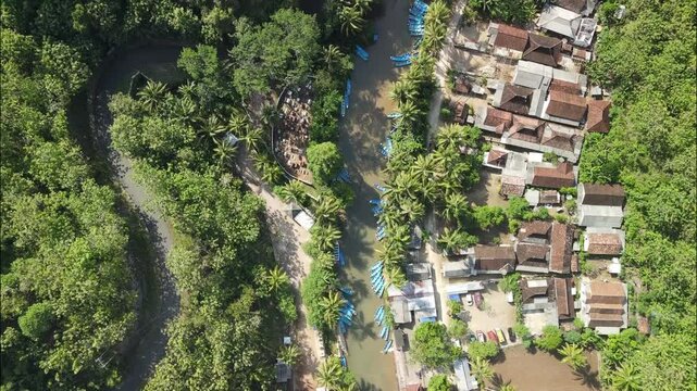 A top-down view reveals a bustling riverside tourism hub, with dozens of blue tour boats lining the banks next to a traditional village and lush jungle. Aerial view Maron river in Pacitan, Indonesia.