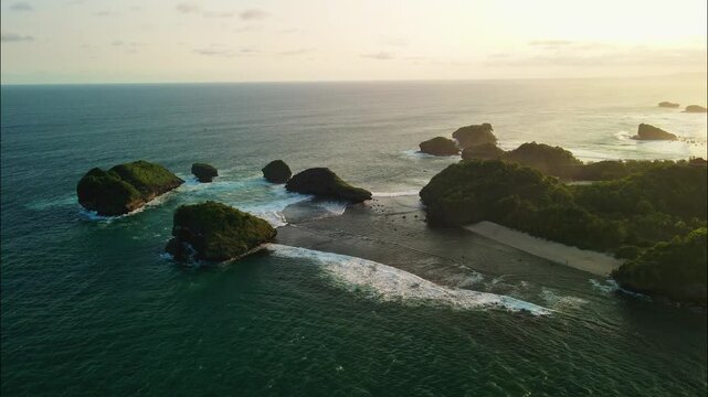 A stunning seascape unfolds with numerous green-topped sea stacks and rocky islands scattered across the ocean, all bathed in the warm, hazy light of sunset. Watu Karung Beach, Pacitan.