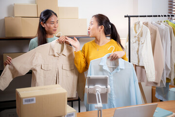 Two young asian woman live online clothing store while showing clothing with tablet on social at...