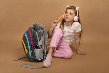 Little schoolgirl sitting next to school backpack in pink headphones and pencil thoughtfully...