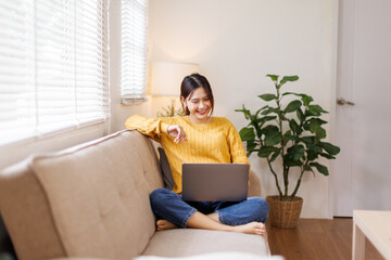 Young Asian woman doing research work for her business. Smiling woman sitting on sofa relaxing...