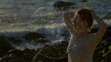 Woman gathering hair on rocky beach, summoning digital globe pulsing glowing network in technology - Powered by Adobe