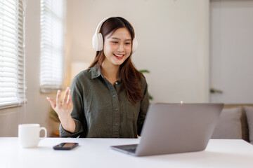 Asian woman wearing headphones and raising one hand while sitting in front of a laptop on white...