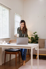 Young minded successful employee business asian woman in blue shirt work hold cup coffee to go stand at desk with laptop pc computer at office indoor Achievement career concept
