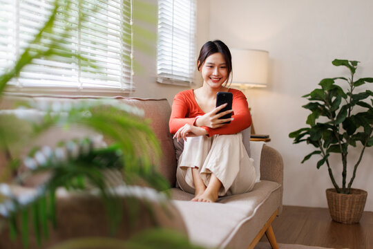 Happy young asian woman relax on comfortable couch at home texting or chatting messaging on the phone, smiling girl use cellphone, online message, shopping online from home
 - Powered by Adobe